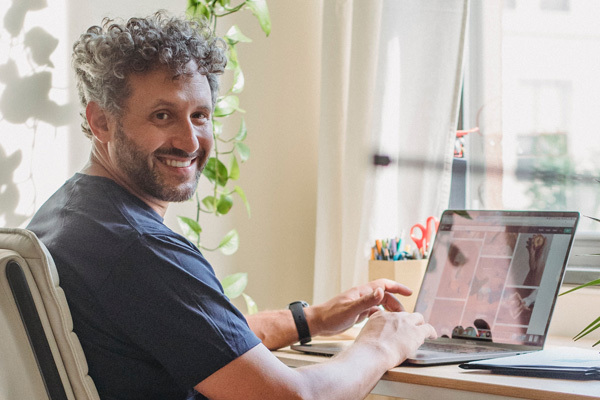 Smiling adult in a bright room facing a window. They are sitting at a desk typing on a laptop.