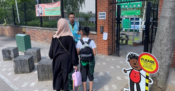 A woman and boy stand in front of a school gate near a sign that says Please turn off your engine