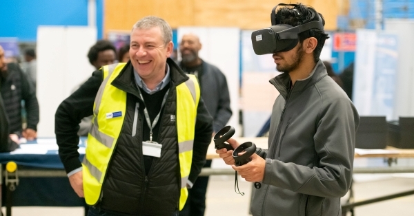 A young man uses a VR headset while another in a high vis jacket looks on