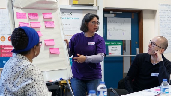 A woman stands by a white board covered in post it notes at the front of a class watched by adult students