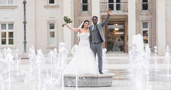 A wedding couple wave as they stand in the middle of the fountains in Fellowship Square