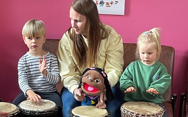 Two young children play bongos either side of an adult holding a glove puppet
