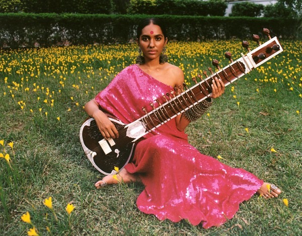 Photo of a woman sitting in a field with a sitar in a pink sequined dress.