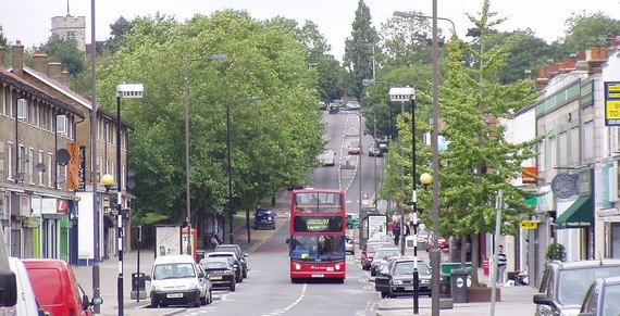 A number 97 bus drives down the hill towards Chingford Mount with trees eitehr side and behind and cards parked along both sides of the road