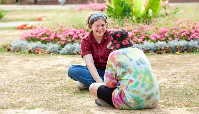 Two young people sit on parched grass in a park on a sunny day