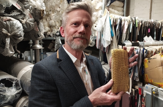 Andrew Wagland holds up a roll of gold braid in front of rolls and rails of fabric