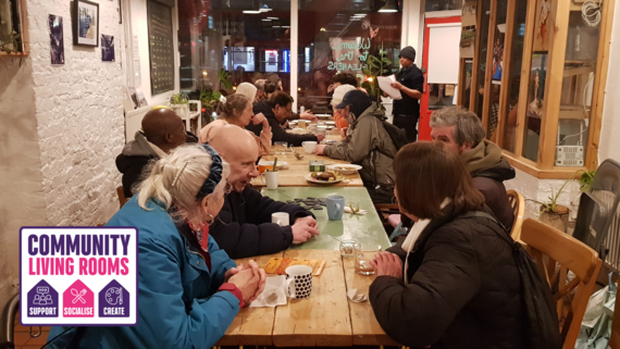 A large group of people socialise around a table in a community living room