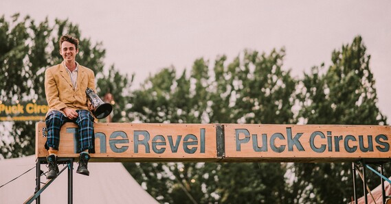 A smiling man with a megaphone in his hand sits on top of a sign that says The Revel Puck Circus