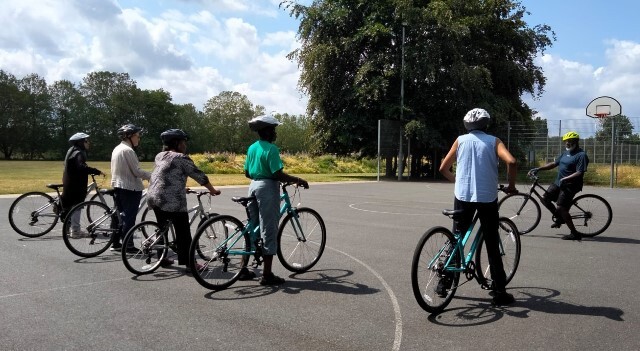 A group of cyclists wearing helmets listen to a trainer on a basketball court in Leyton Jubilee Park
