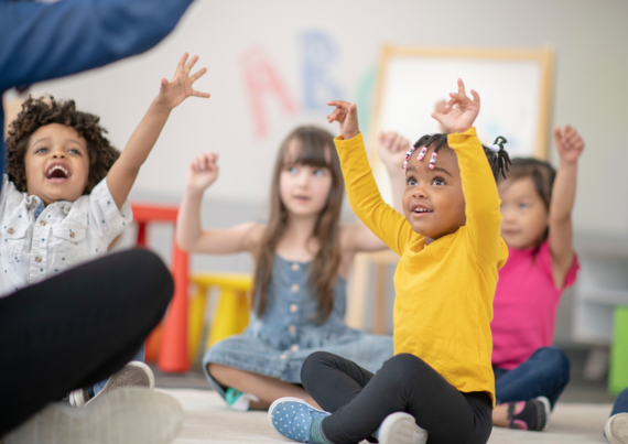 Pre-school children sit on the floor with the arms raised and their mouths open in song
