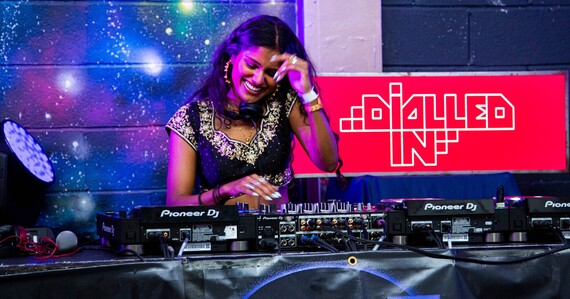 A smiling DJ uses decks in front of a Dialled In poster 
