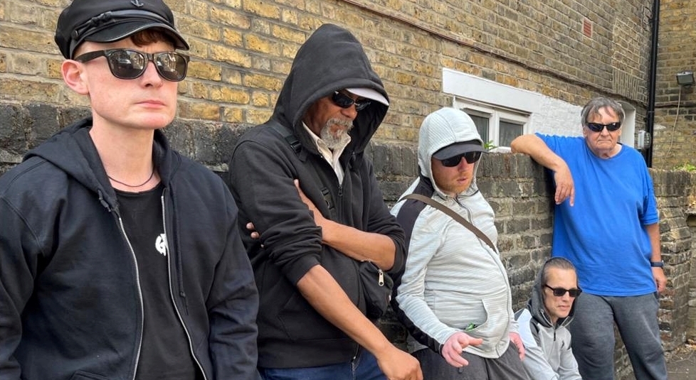 A group of men in dark glasses lean against a brick wall