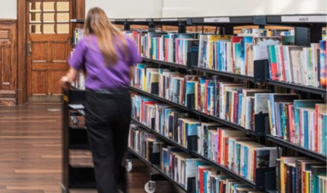 A blurred figure is pictured from behind pushing a trolley as they walk beside library shelves of books