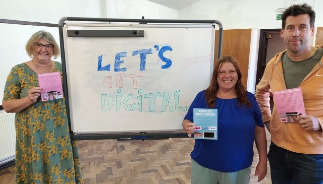 Three people holding up leaflets pose by a flip chart. It has Let's get digital written on it