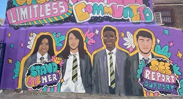 A mural on a wall shows four school students in uniform, including the words Stand by her, See report support, limitless and community