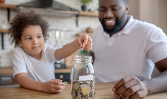 A father and child sit at a kitchen table. The child puts a coin in a jar full of cash.