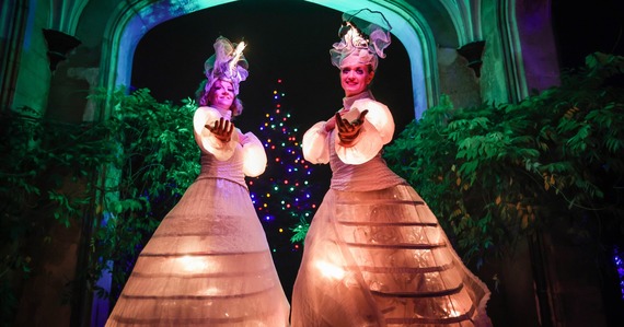 Two performers in illuminated crinoline skirts and puffed sleeves stand in front of an illuminated tree and other foliage