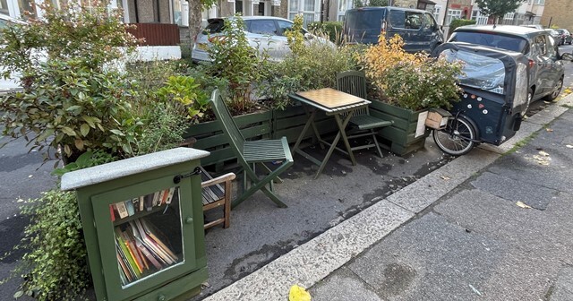 A parklet in a residential street featuring a cabinet full of books, a table and chairs and greenery 