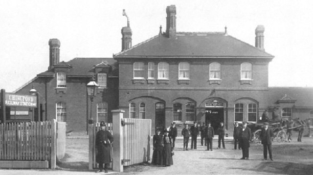 Station workers in uniform, a police office, women in hats and long skirts and a horse and cart stand outside Chingford Station in the late 1800s