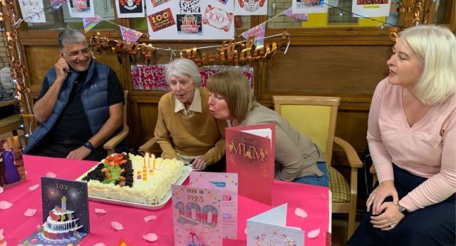 A woman helps Louise blow out the candles on her birthday cake while others look on. There are birthday cards in front of the cake