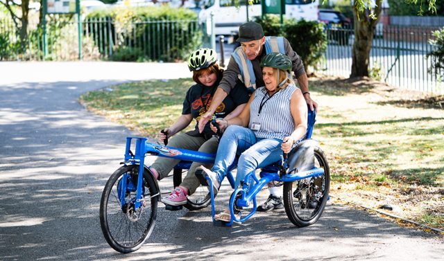 Two people sit in a double bike in a park as another person shows them how to use it