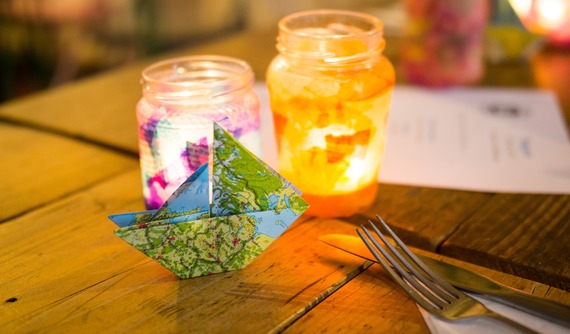 Close up on an origami boats with candles in jam jars behind and a knife and fork to the side