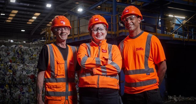 Three people in high vis jackets and hard hats stand in front of a huge pile of recycling