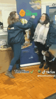 Two people from The Hive wearing warm jackets and walking boots high five while a dog looks on