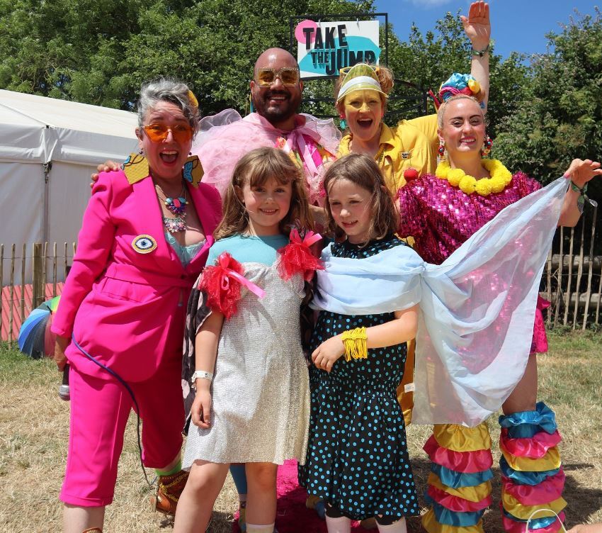 A group of people in brightly coloured costumes in front of a take the jump sign and a tent