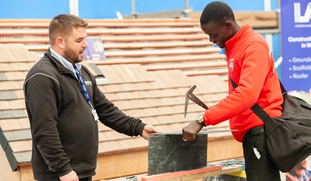 A young man tries out a new skill as an older man looks on at a green construction fair