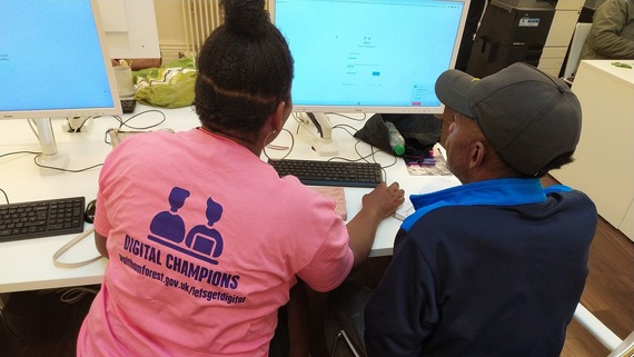 A man and woman are shot from behind. The woman is wearing a Digital Champions T-shirt and showing the man how to use a computer