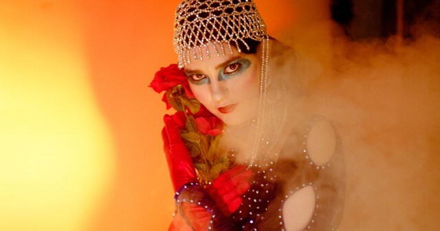 Close up on a person wearing heavy make-up, gloves and a sparkly beanie hat clutching red roses to their face and chest 