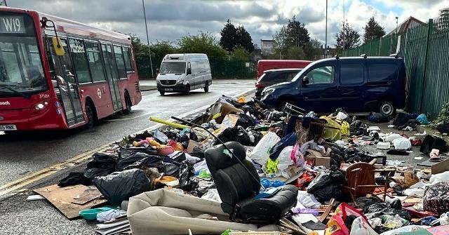 A W19 bus and a white van drive by a huge pile of rubbish dumped on the side of the road