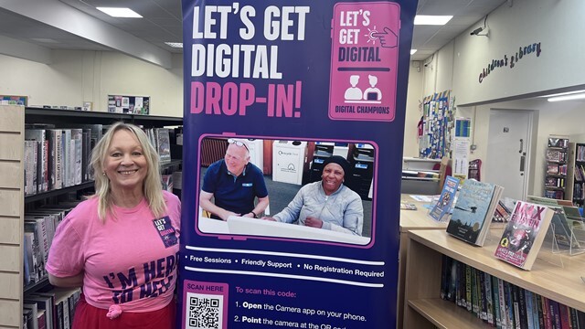 A woman in a pink T-shirt stands by a Let's Get Digital poster in Chingford Library