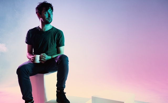 Nathan Fakes sits on a white cube against multicoloured lighting with a mug in his hand.