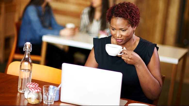 A woman sits at a table in a cafe, drinking a coffee while looking at a laptop
