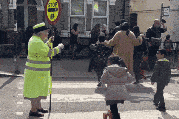 A lollipop lady sees children across the road on the way to school. One child waves as she crosses the road on her scooter