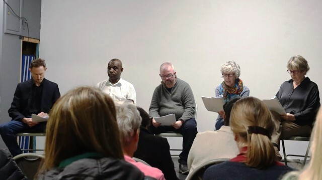 A group of performers sit on chairs reading from scripts in front of an audience