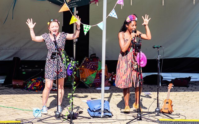 Two women wearing summer dresses and flowery headresses perform on stage