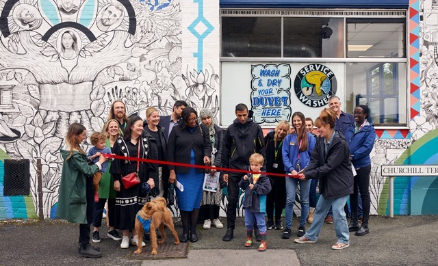 A child cuts a red ribbon in front of a crowd and a dog, in front of the Chingford Mount mural