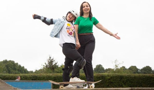 A woman poses with a child on a skateboard with their arms spread wide. They are smiling.