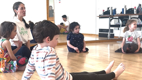 A woman sits with young children on the floor teaching a dance class