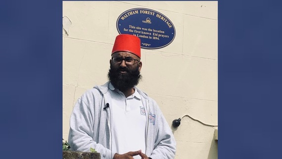 A photo of a man speaking in front of a blue plaque