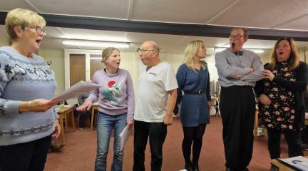 Six actors standing in a line holding scripts rehearsing a pantomime