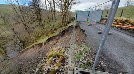 Picture showing landslip at Cowgill, with fencing around washed away section of road and steep drop to a stream below