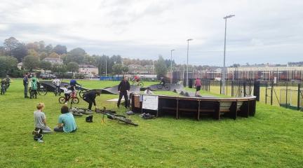 A busy pump track with groups of young people sitting on the grass and people using it