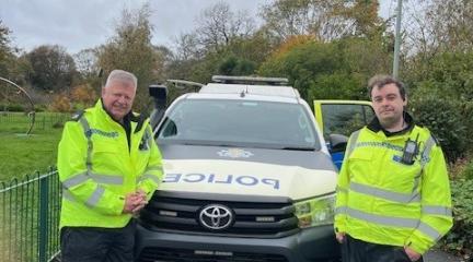 Two CCTV operators in hi-vis yellow jackets standing beside a police car in a park. CCTV camera in the background