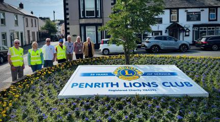 A group of people standing beside a display of yellow and blue flowers with a Penrith Lions Club banner at the centre