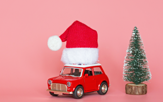 A red car with a santa hat on the top and a christmas tree standing next to it