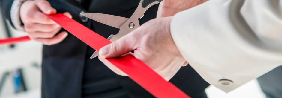 close up shot of a man's hands holding a red rbbon while a woman's hands cut it with a pair of scissors